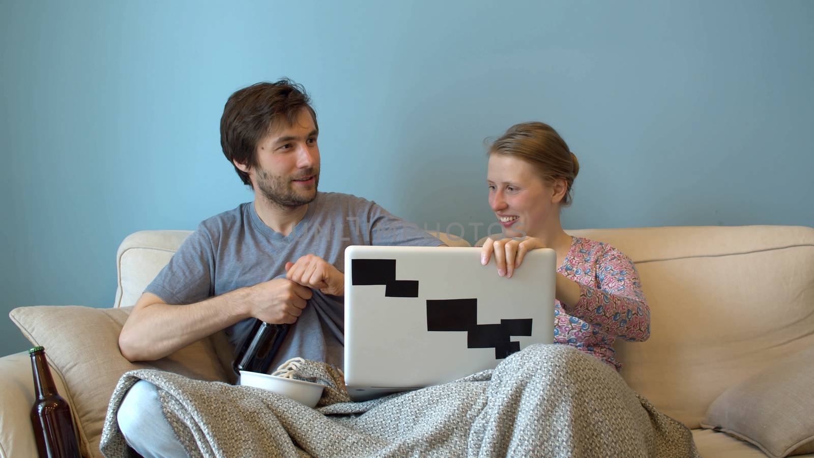 Man opening bottle of beer. Family sitting on the couch and watching a movie or broadcast on a computer. Happy couple in a cozy room on the sofa under the plaid