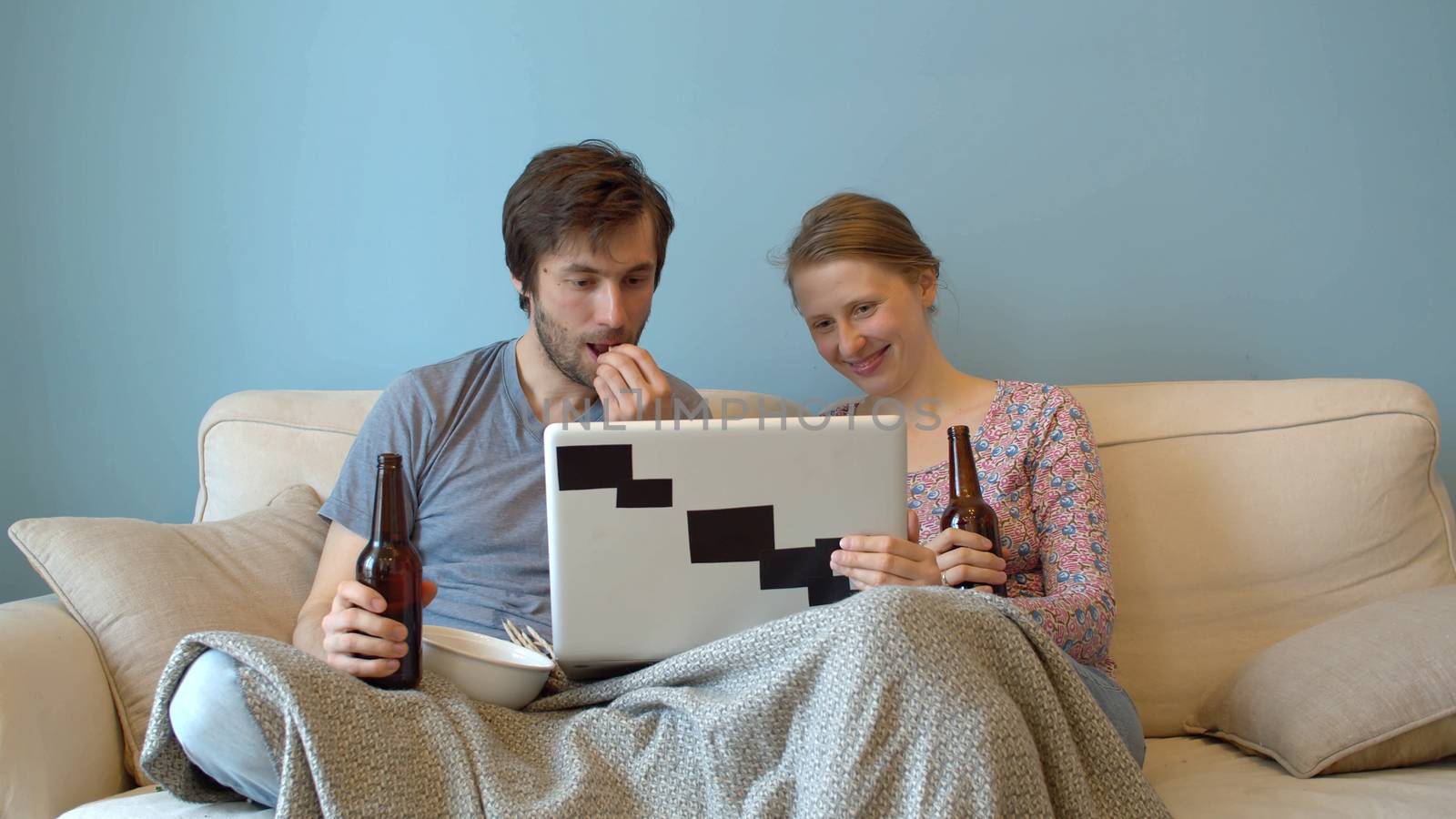 Family drinking beer on the couch and watching a movie or broadcast on a computer. Happy couple in a cozy room on the sofa under the plaid