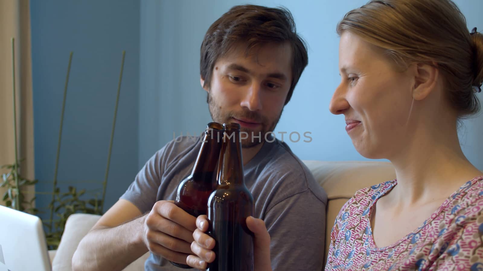 Close up couple clinks beer bottles. Family drinking beer on the couch. Happy couple in a room on the sofa