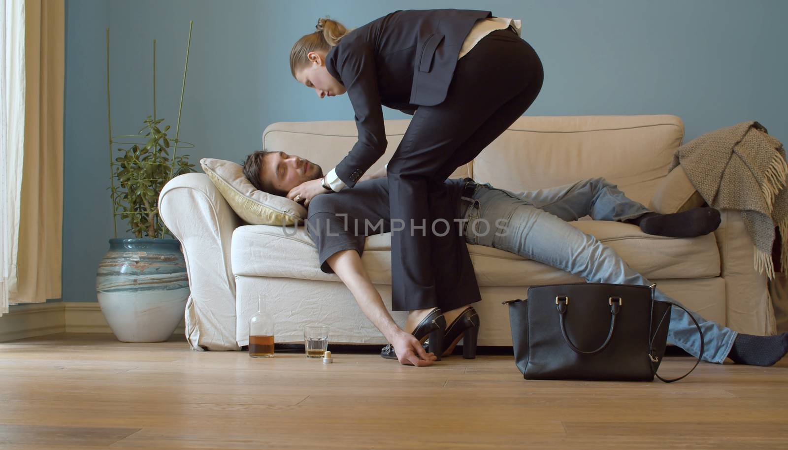 Drunk man sleeping on the couch dressed. His wife in a business suit take care of him. Young debauchee in his apartment after party