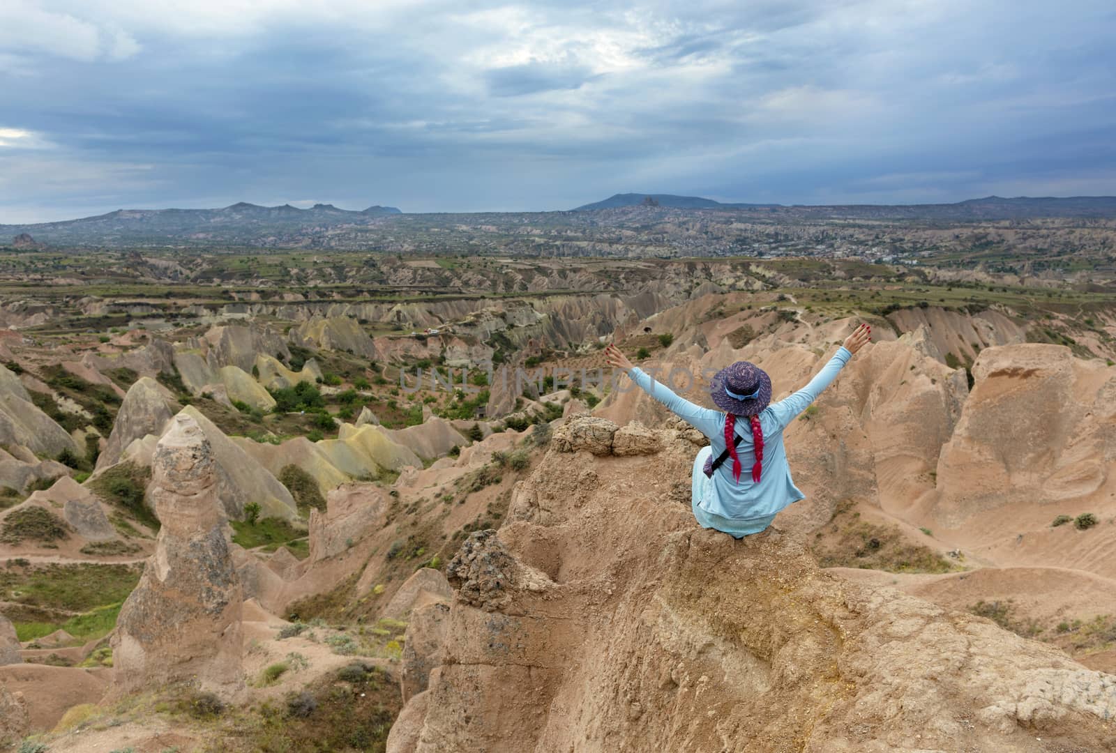 A young girl in turquoise clothes sits on top of a rock in Cappadocia and looks spread her arms out on mountains, ravines and a blue sky.