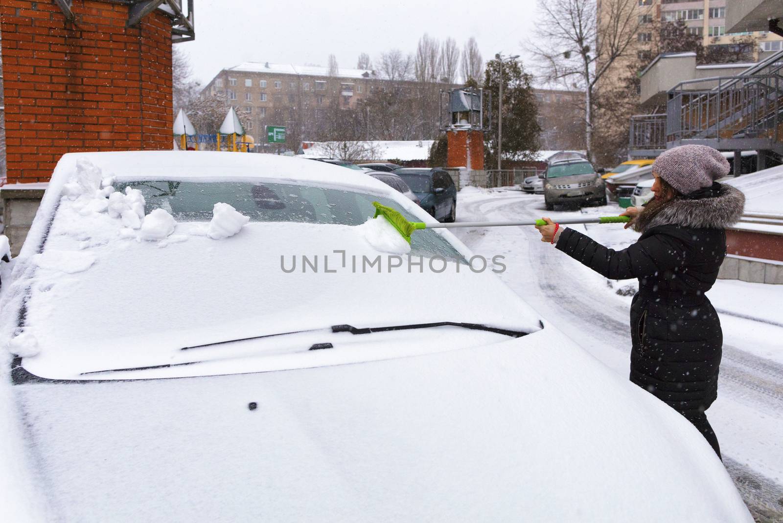 A young woman cleans snow from the surface of her car. by Sergii