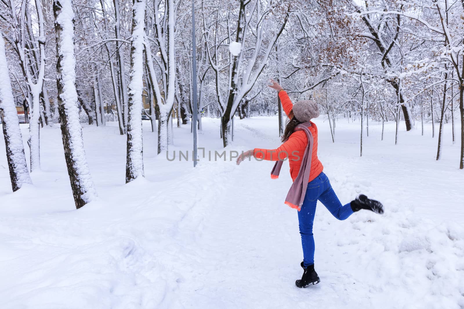 A young beautiful woman in a bright coral jacket and blue jeans in winter walks along the alley of the snow-covered fairy-tale park and throws a large snowball.