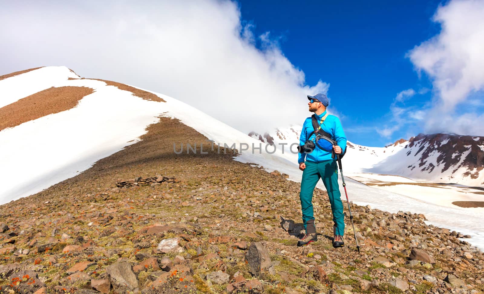 Tourist photographer with a cane in his hand stands on the slope of a snow-capped mountain peering at its top and planning to climb against the background of a bright blue and cloudy sky