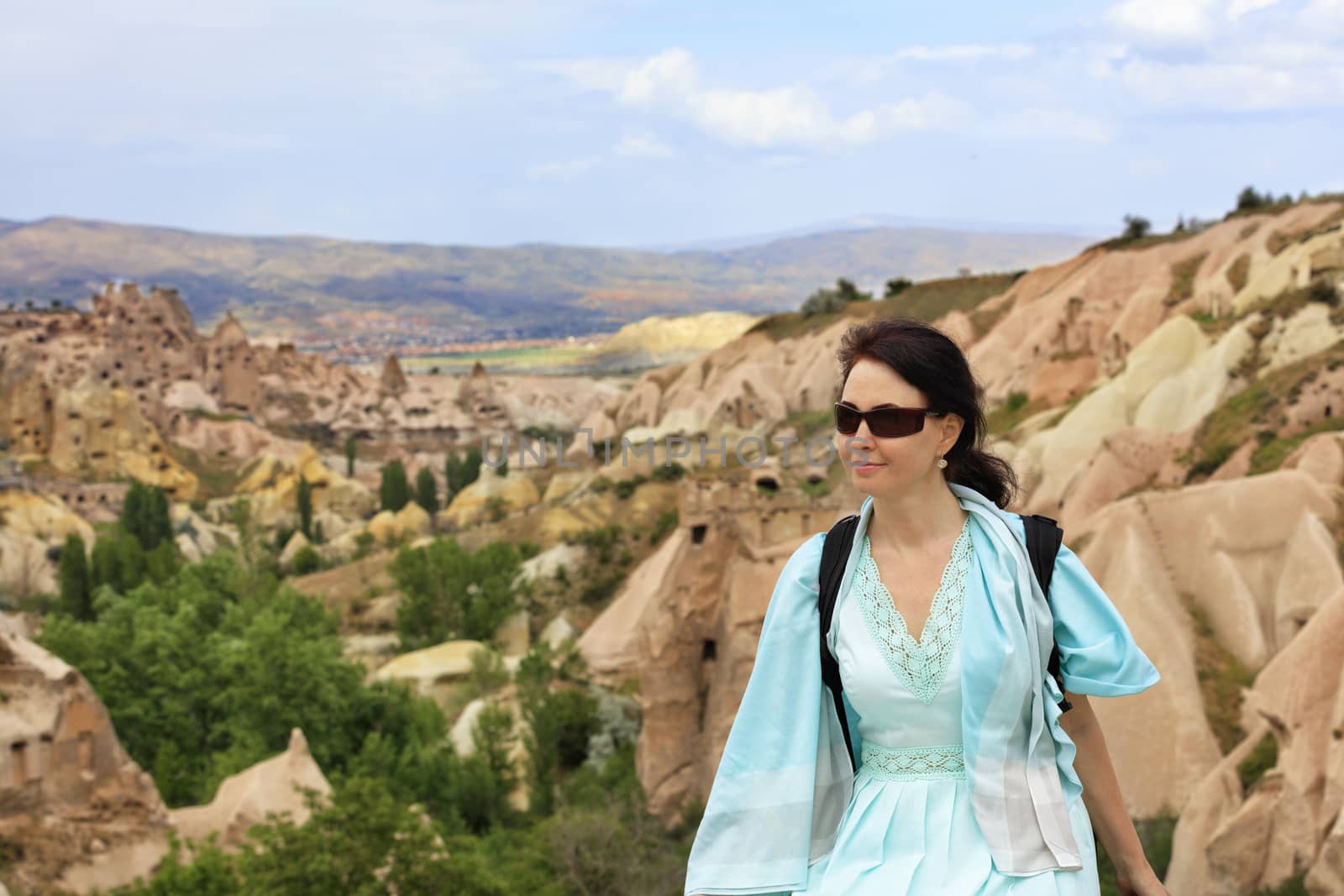 Portrait of a young beautiful woman in sunglasses on her face, a turquoise dress and a backpack on her shoulders against the blurred landscape of mountain caves in the valleys and canyons of Cappadocia