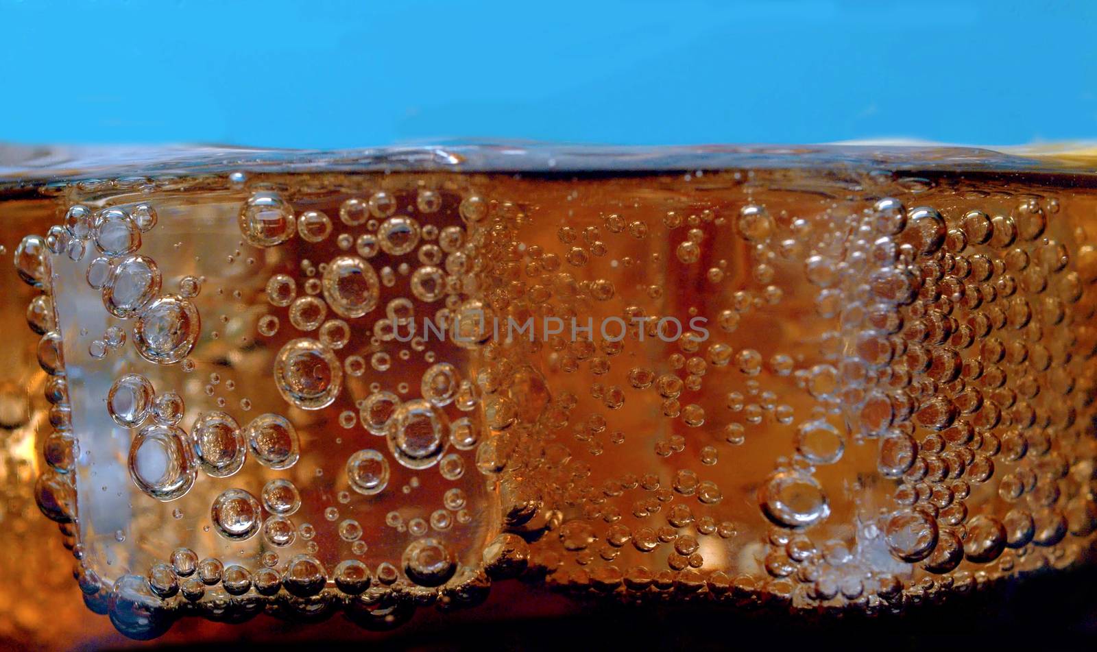 Rising gas bubbles in a glass with cola extreme close up. Pieces of ice floating on the coca surface on blue background. Soda carbonated drink into a drinking glass