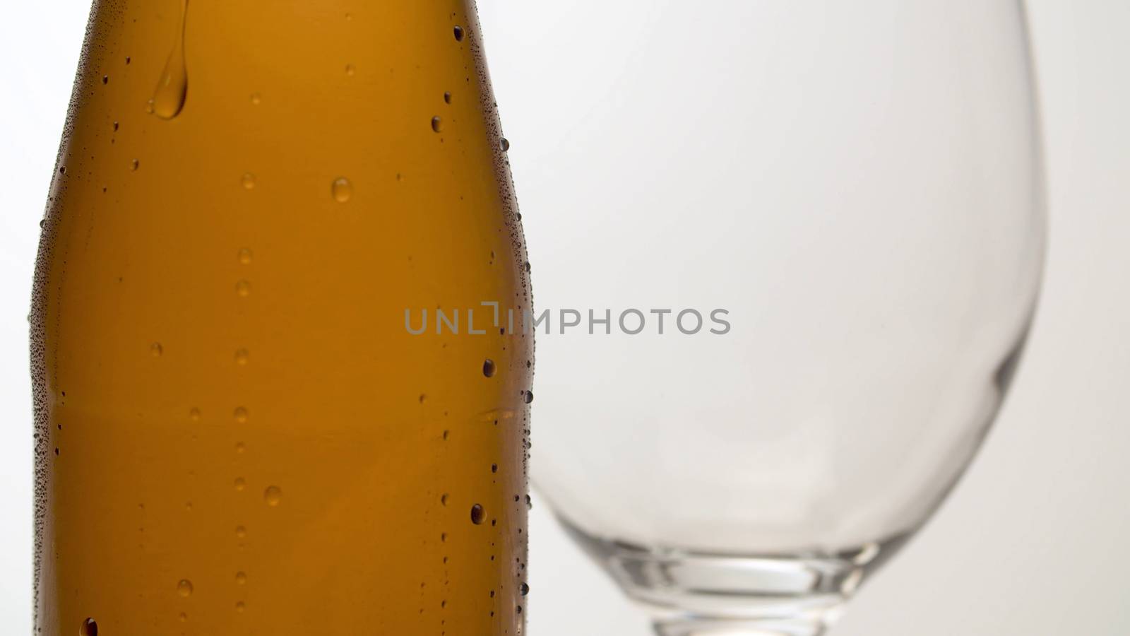 Close up misted bottle of cold beer on the table. Wineglass behind it. Macro shot on white background. Drops of water flowing down. Craft beer closeup