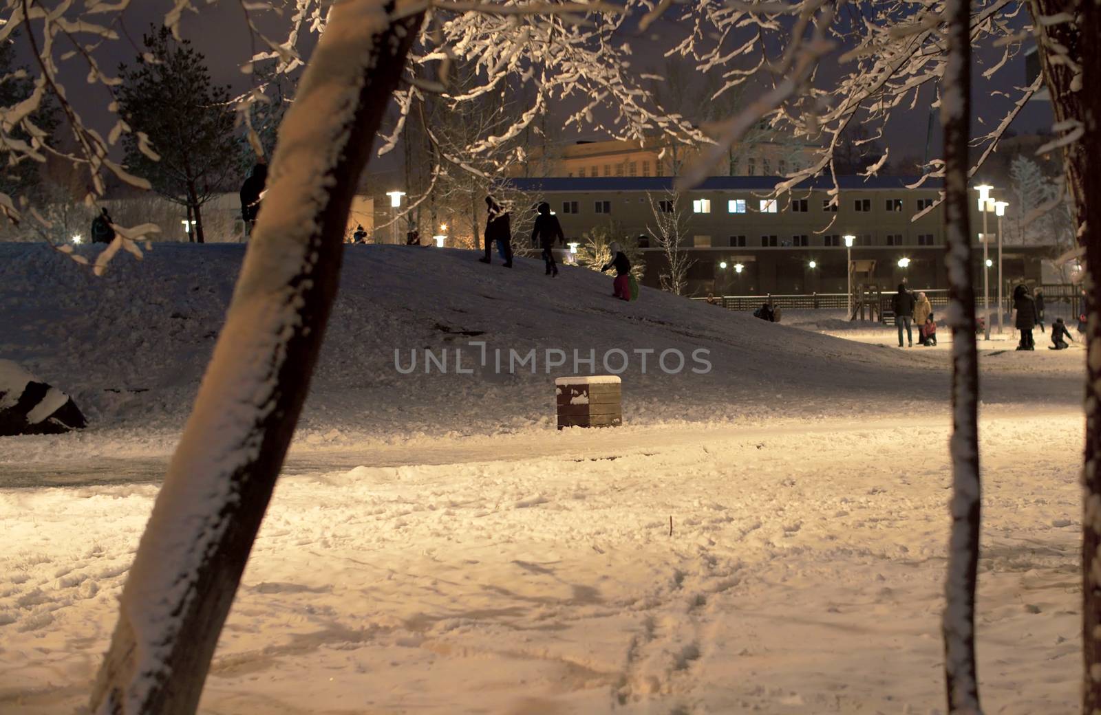 Snow covered city park in the evening. Light of lanterns. Winter cityscape. Children walking on the kid's playground