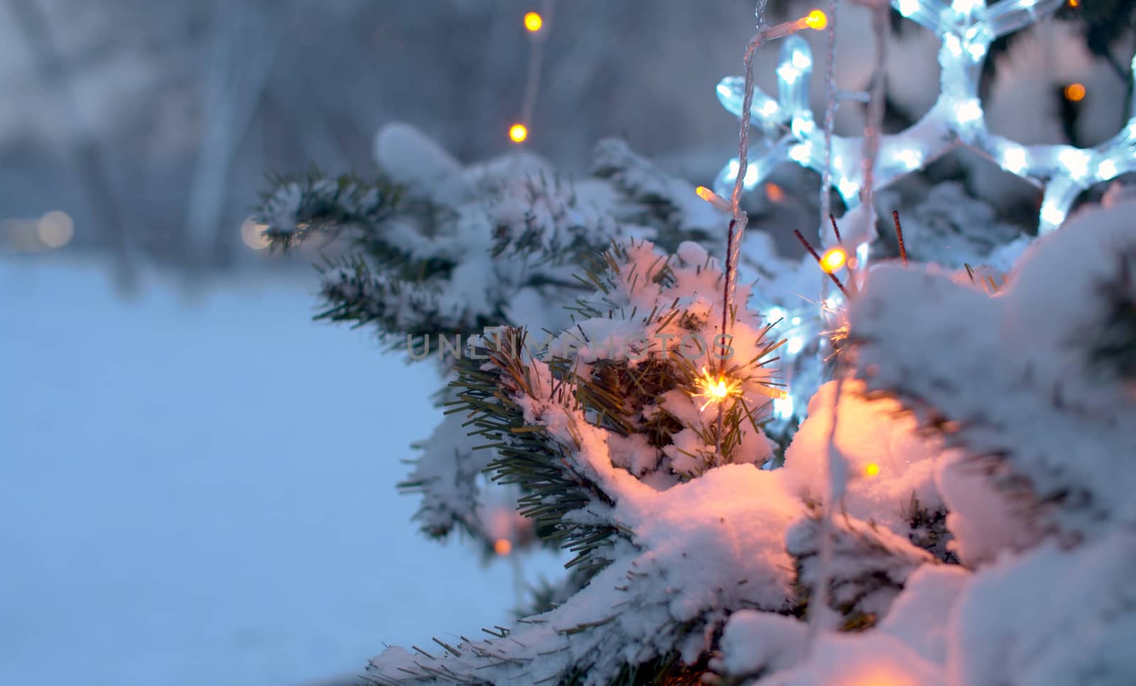 Close up sparklers burning on a Christmas tree in a park. Blurry lights of a garland. City holiday decorations in the evening. Snowfall in the city.