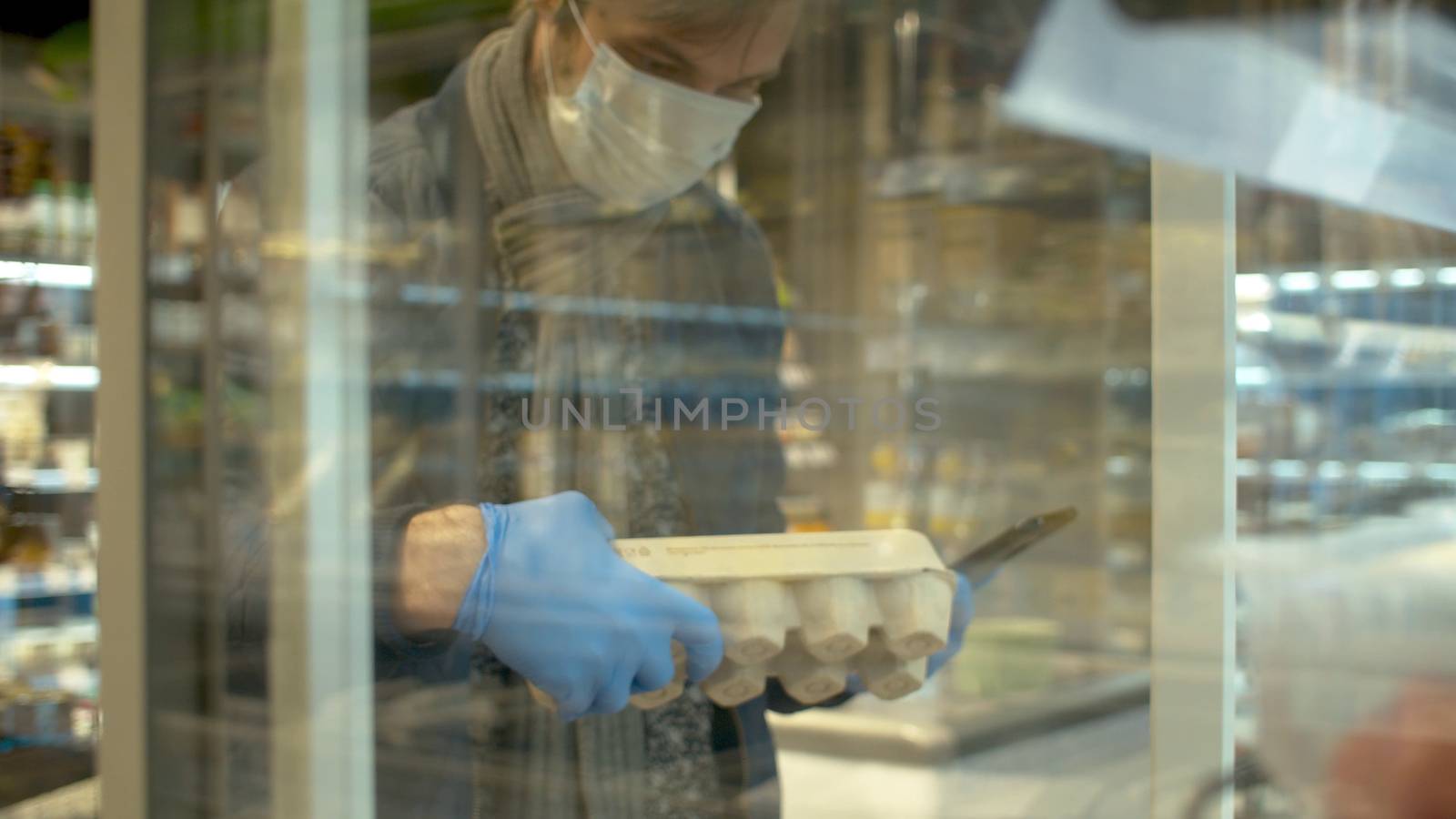Male portrait through the glass door. Young handsome man in a protective mask and gloves choosing eggs in the supermarket. Coronavirus epidemic in the city.