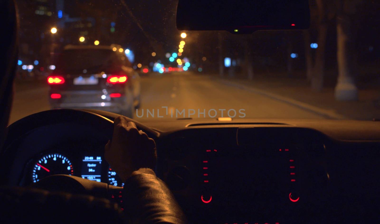 Close up man's hands on the steering wheel of a car. Console and instrument panel lights up inside the dark interior of a car while driving at night. POV. Blurred lights on the city street
