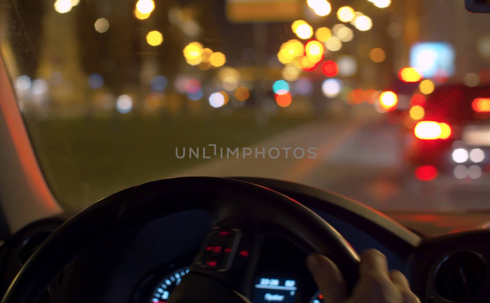 Blurred traffic lights in the city. Close up man's hands on the steering wheel. Dashboard lights up inside the dark interior of a car. POV night driving.