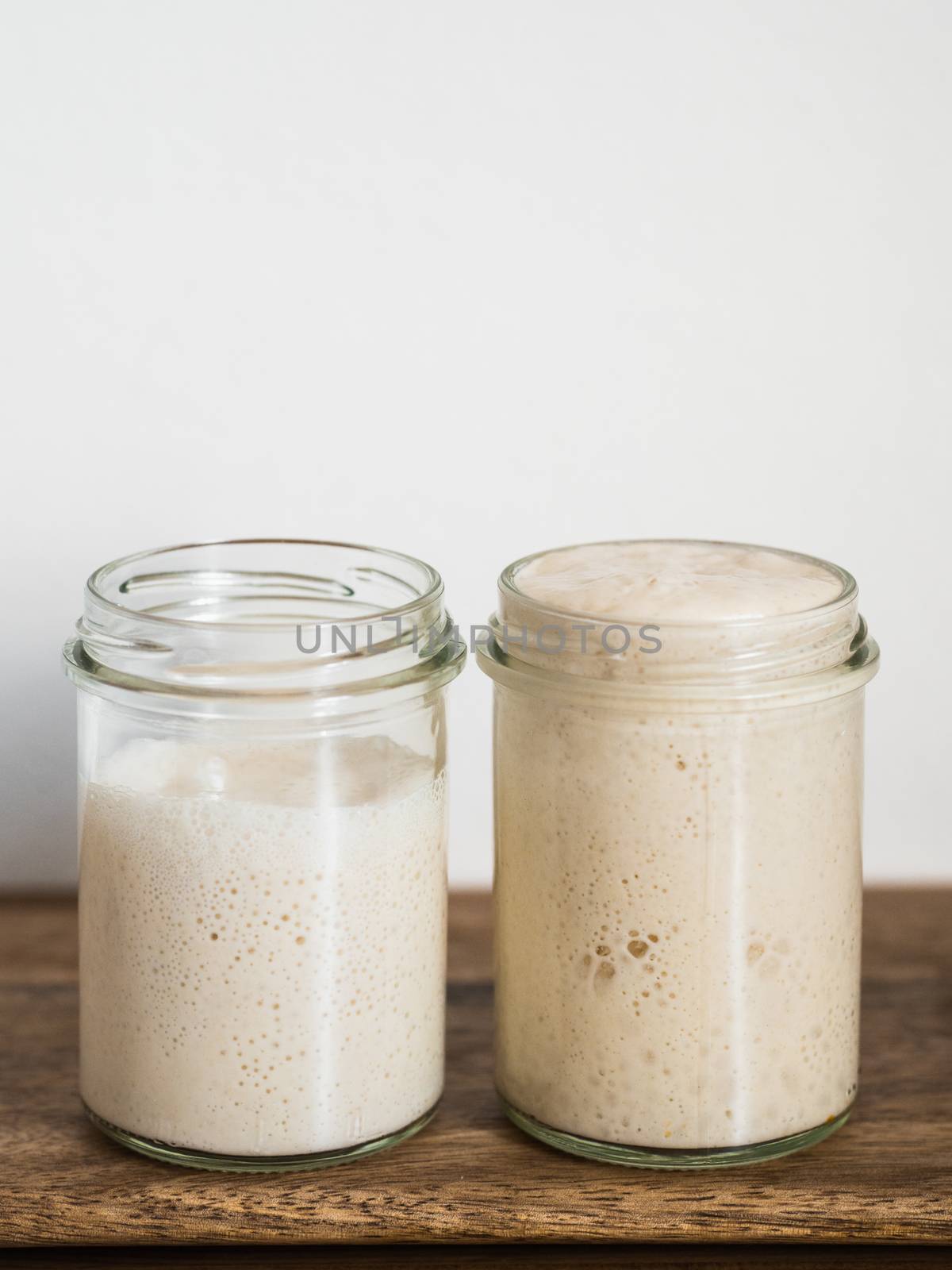 Two glass jars with wheat sourdough starters in different hydration levels. Starter 100 percent hydration (right) and starter with higher hydration (left). Copy space top. Vertical.