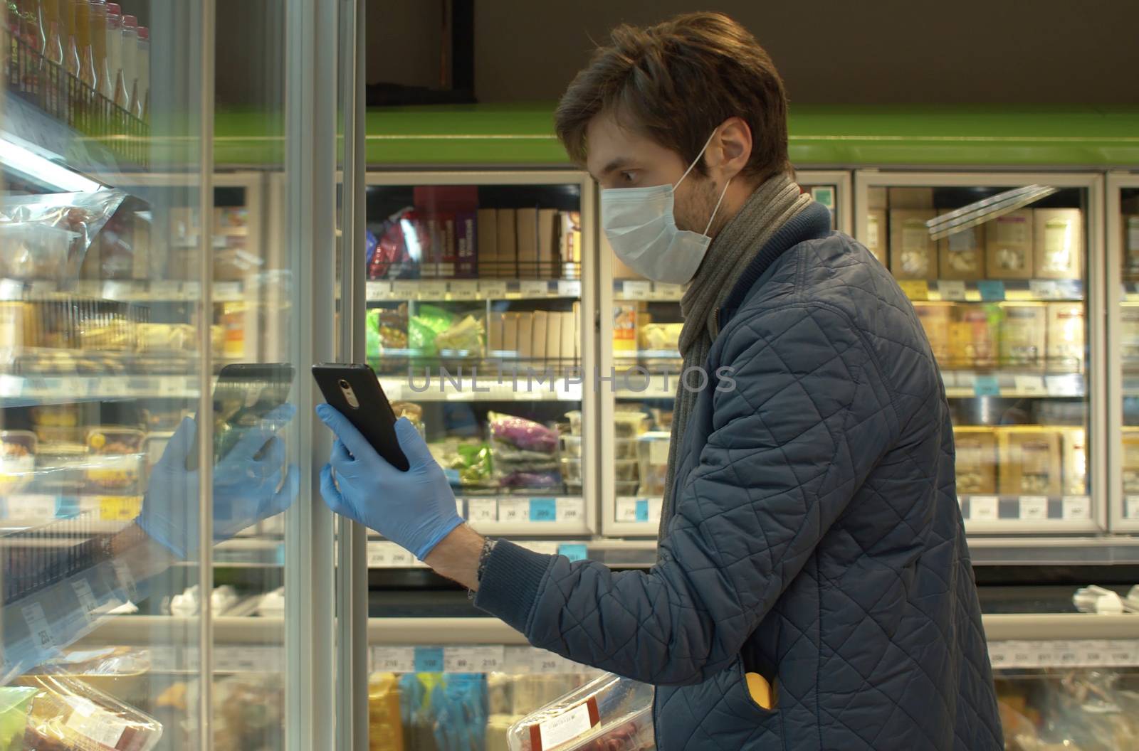 Young handsome man in a protective mask and gloves in the supermarket. He takes the products from the refrigerator. Coronavirus epidemic in the city.