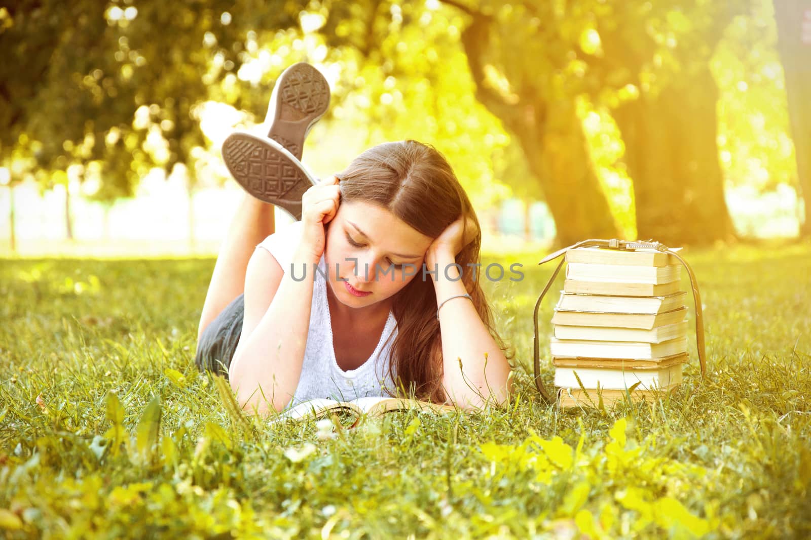 Young beautiful college student girl lying down on the green grass and reading a book at campus at warm day. Education. Back to school conceptual image.