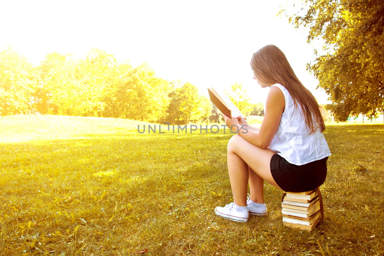 Attractive female college student reading a book and sitting on the stack of books. Education. Back to school conceptual image.