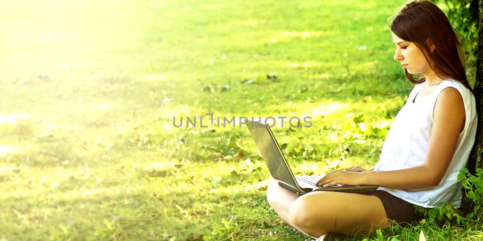 Young beautiful college student girl sitting on the green grass and working on laptop at campus at warm day. Education.