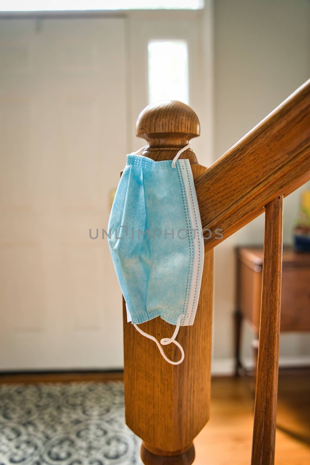 Mask hanging on stair railing looking towards entrance of home. Symbolic of need to wear face mask when leaving home for protecting from COVID-19