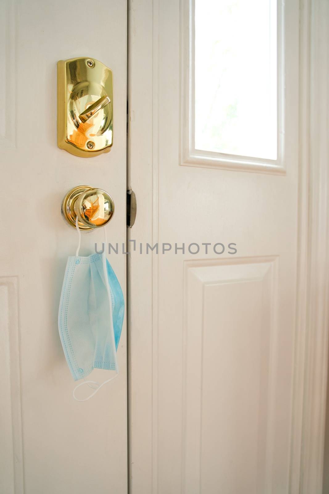 Facemask hanging from door handle on white door with sunlight coming in through window.