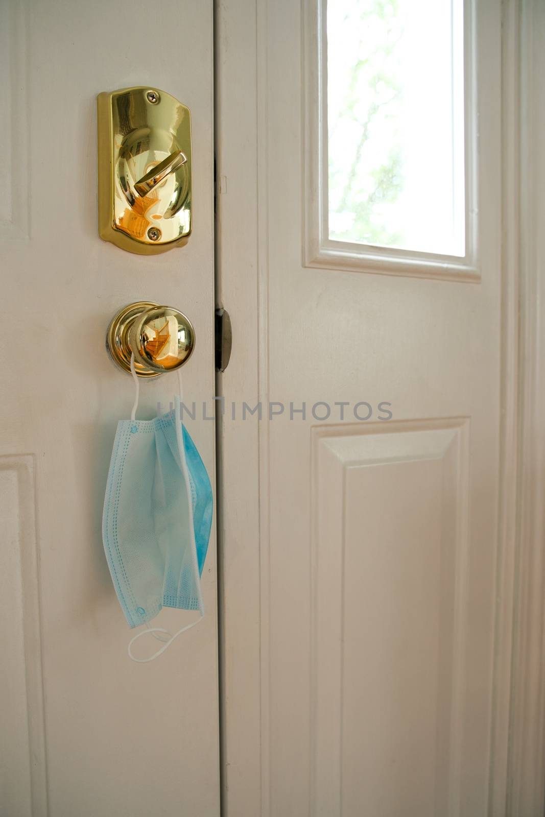 Face mask hanging from door handle on white door with sunlight coming in through window. Covid-19 coronavirus concept image with room for text or copy