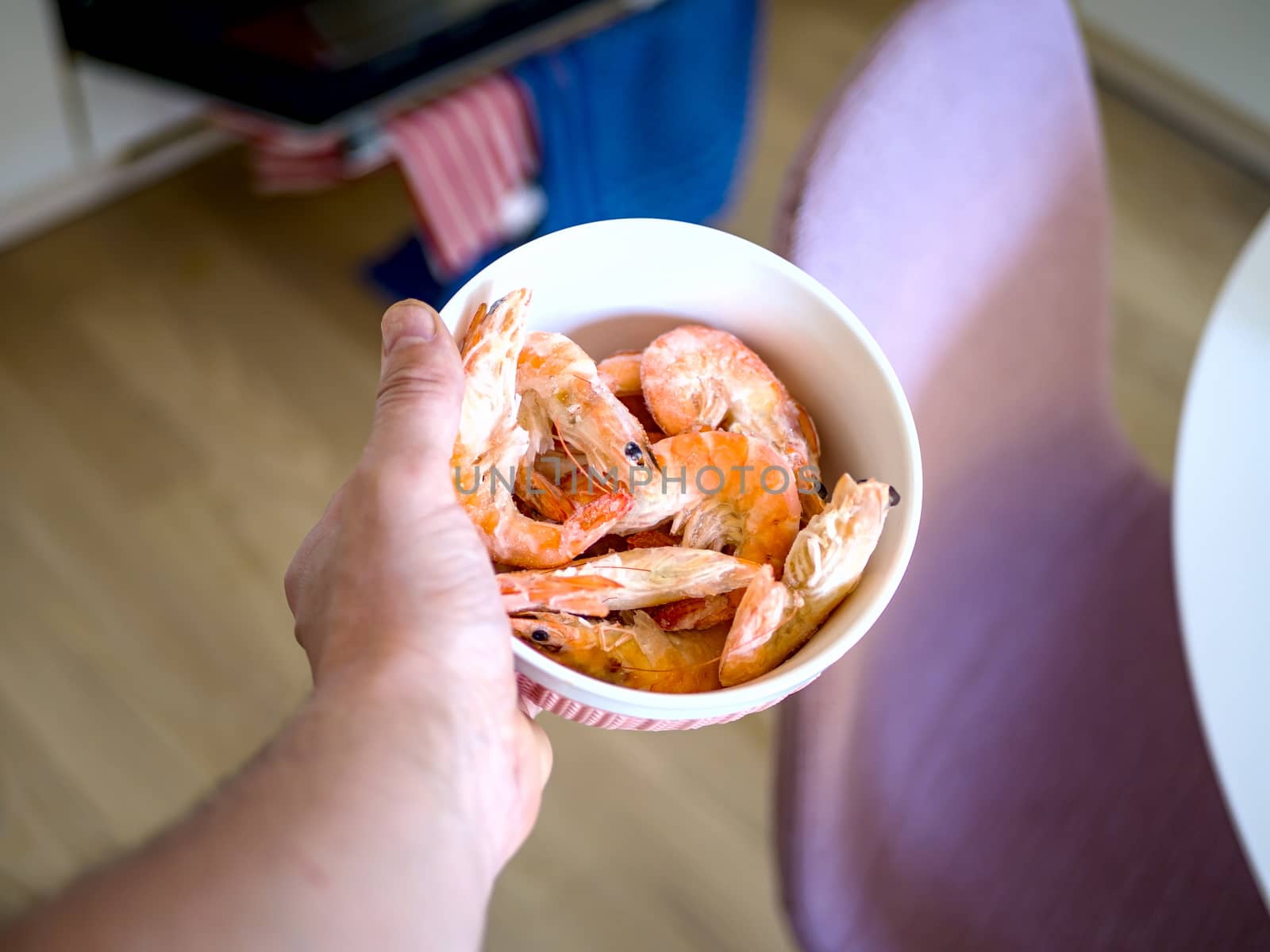 Bowl filled with fresh shrimps in man hands. Chairs are visible in the blurred background. Food for diet - Grilled shrimps with mango salad. Home cook in Asian kitchen.