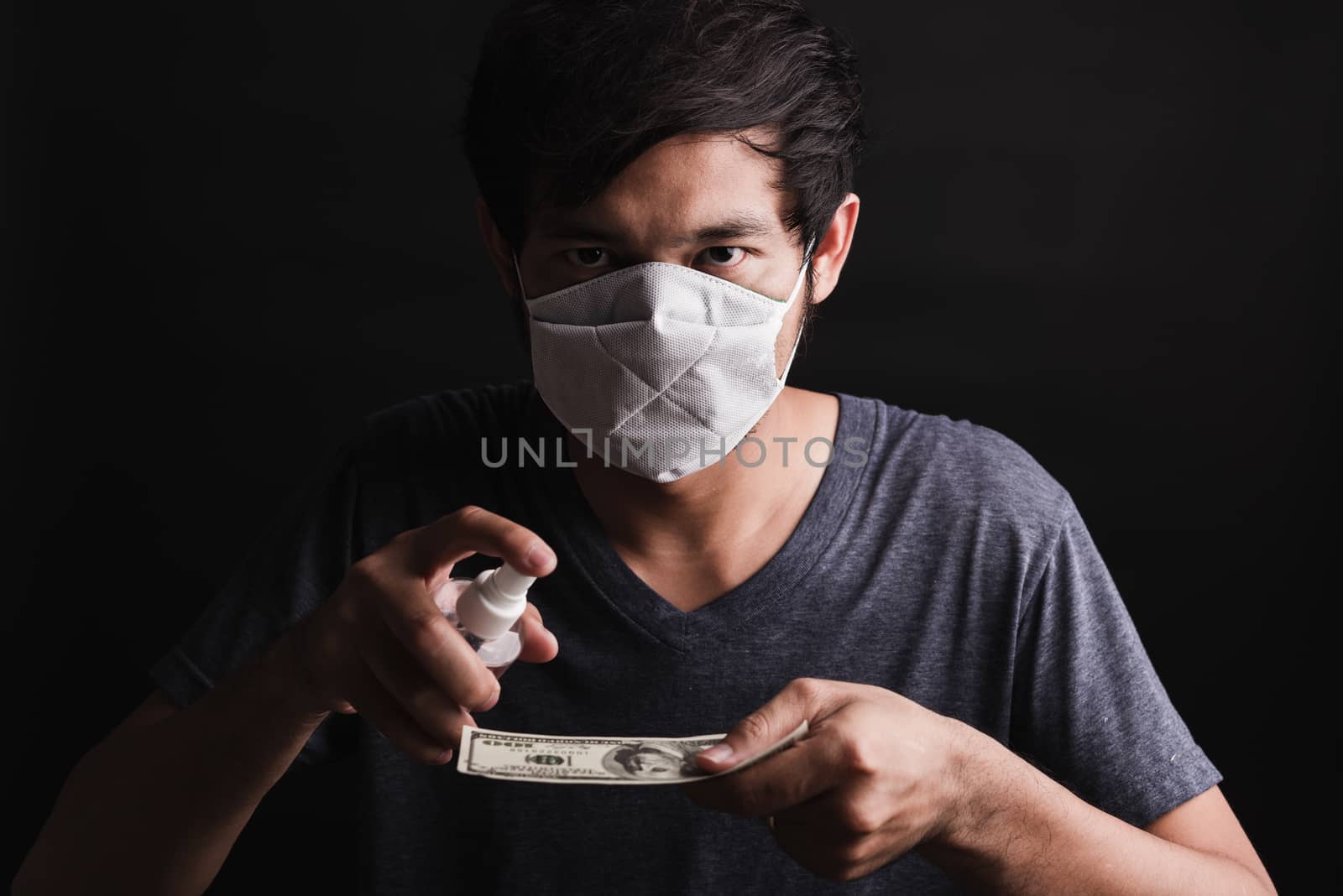 Closeup man wearing protective face mask applying sanitizer spray to 100 dollar money for cleaning, hygiene prevention COVID-19 virus or coronavirus protection concept, dark on black background