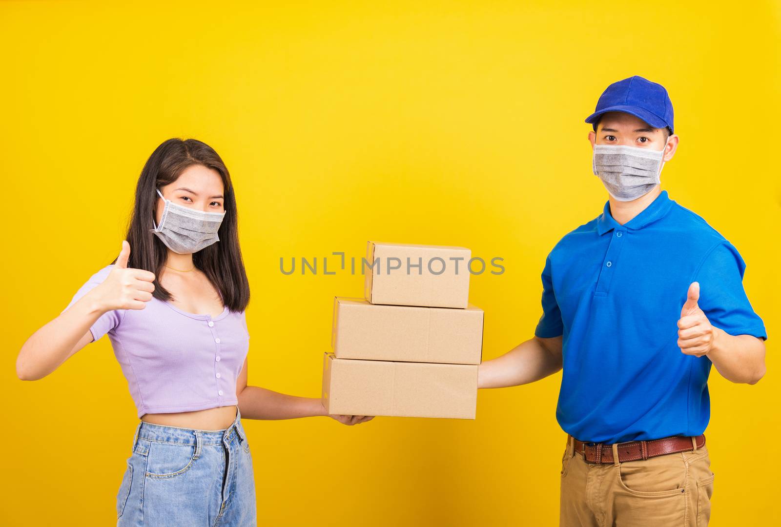 Asian beautiful woman and handsome man wearing protection face mask against coronavirus, accepting delivery boxes, studio shot isolated on yellow background, COVID-19 or corona virus concept