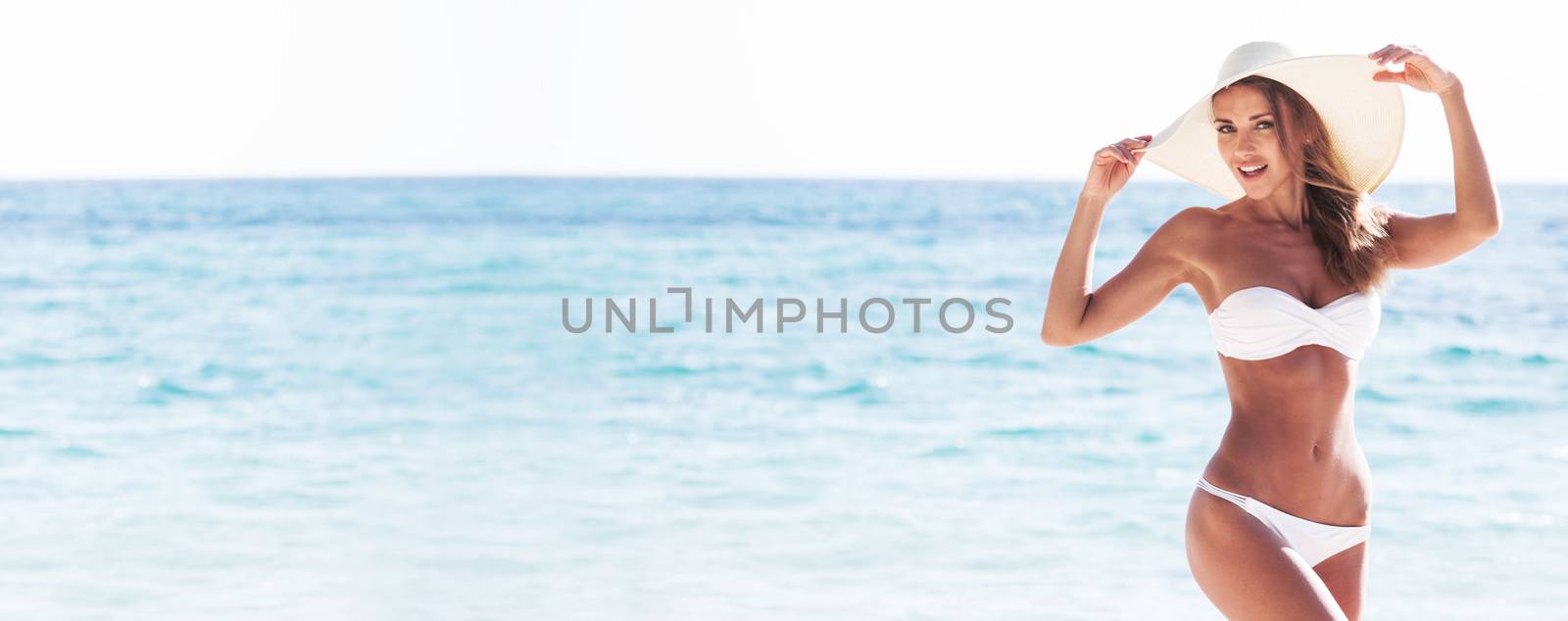 Smiling beautiful woman in white bikini and sun hat at sea beach