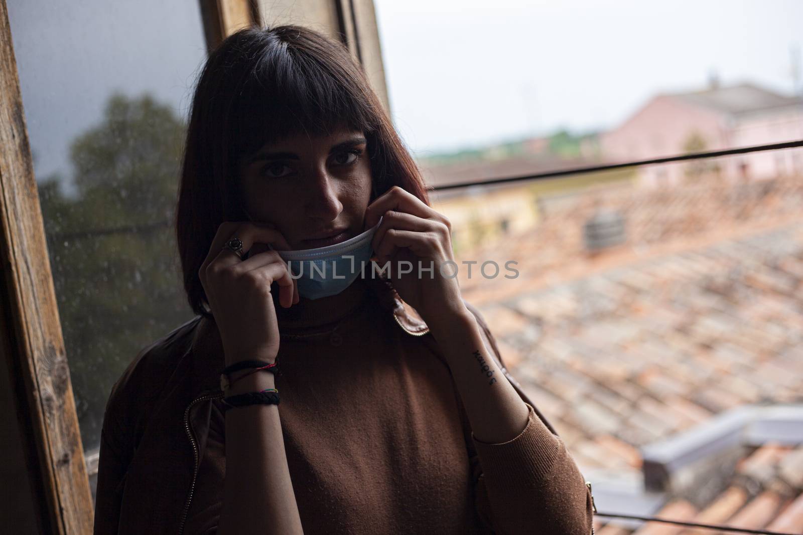 Girl with medical mask at window in her home during covid quarantine period