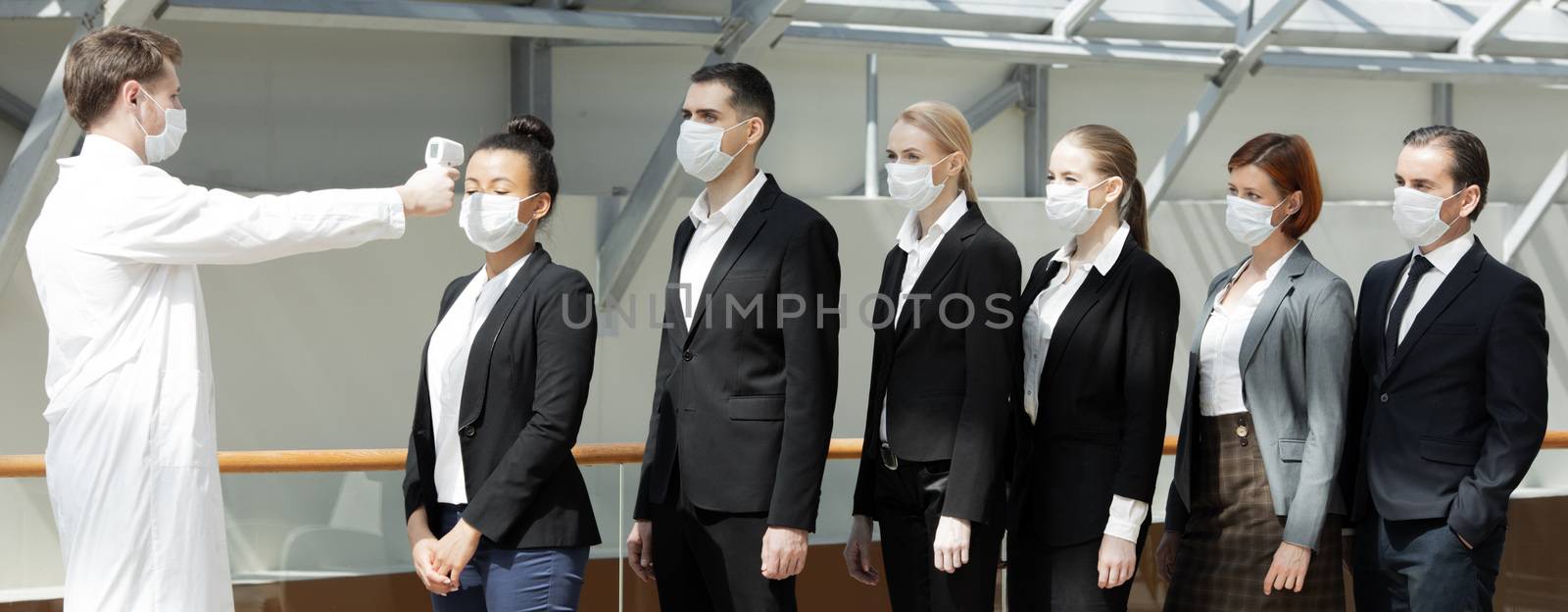 Doctor wearing protective surgical mask using infrared forehead thermometer (thermometer gun) to check body temperature for virus symptoms of business team waiting in a line