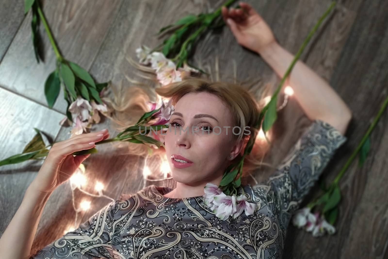 Portrait of a beautiful fashionable middle-aged woman with long blond hair, lying among white flowers and a garland of lights.