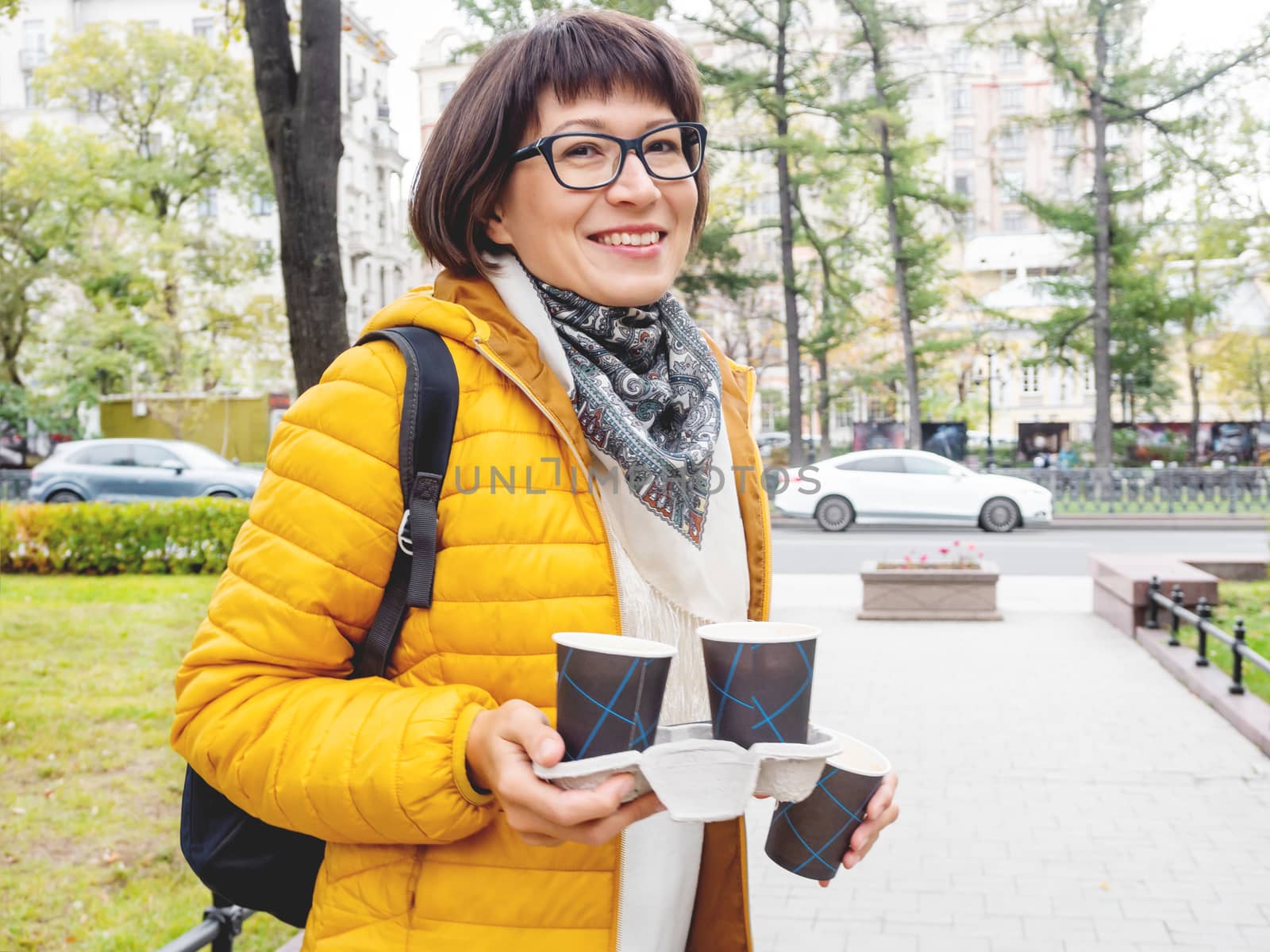 Take away coffee. Happy wide smiling women in bright yellow jacket holds paper cup with freshly brewed cappuccino. Hot beverage on cool autumn day.