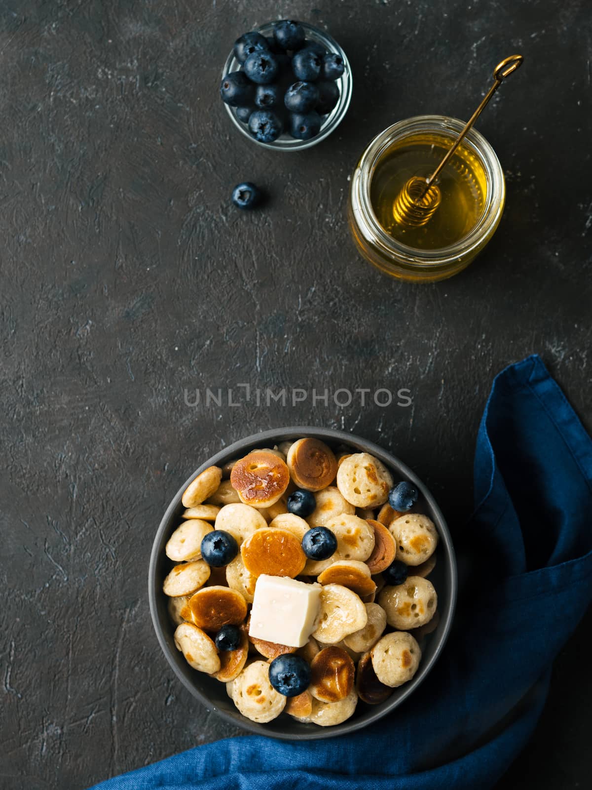 Trendy food - pancakes cereal served butter and blueberries. Heap of mini cereal pancakes in boul on dark background. Copy space right for text. Top view or flat lay. Vertical.