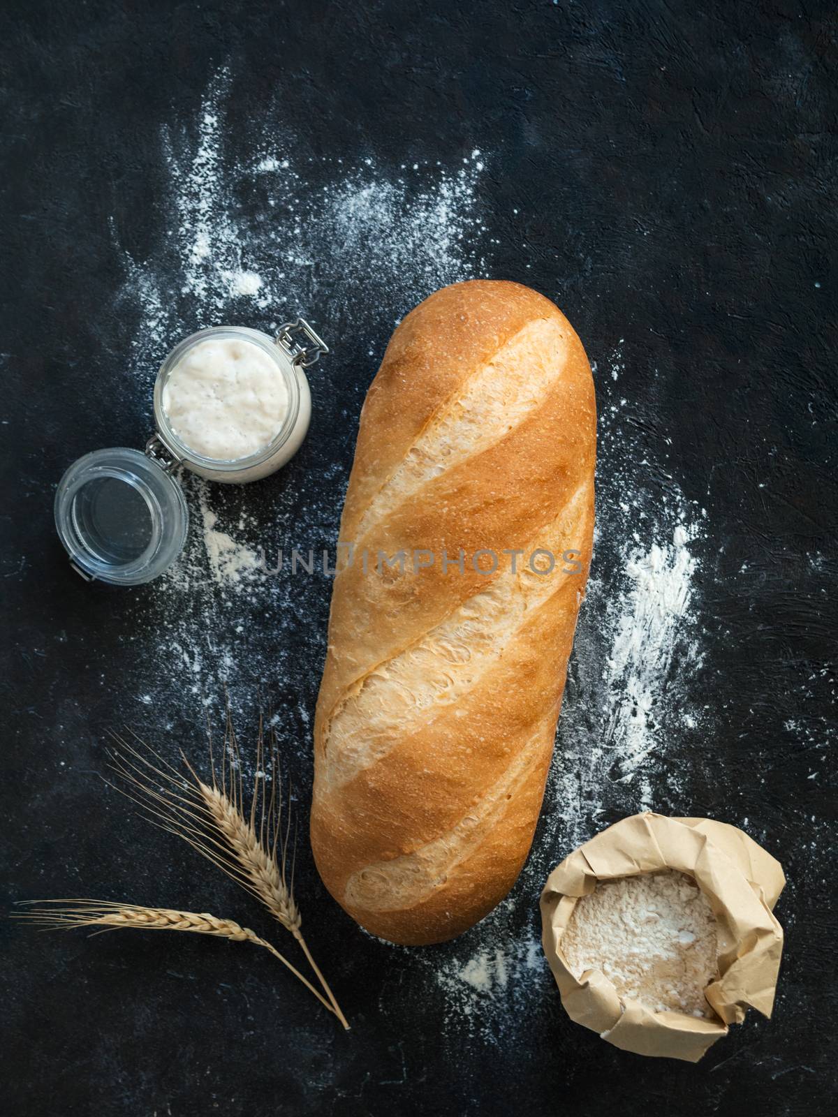 British White Bloomer or European sourdough Baton loaf bread on black background. Fresh loaf bread and glass jar with sourdough starter, floer in paper bag and ears. Top view. Copy space. Vertical.