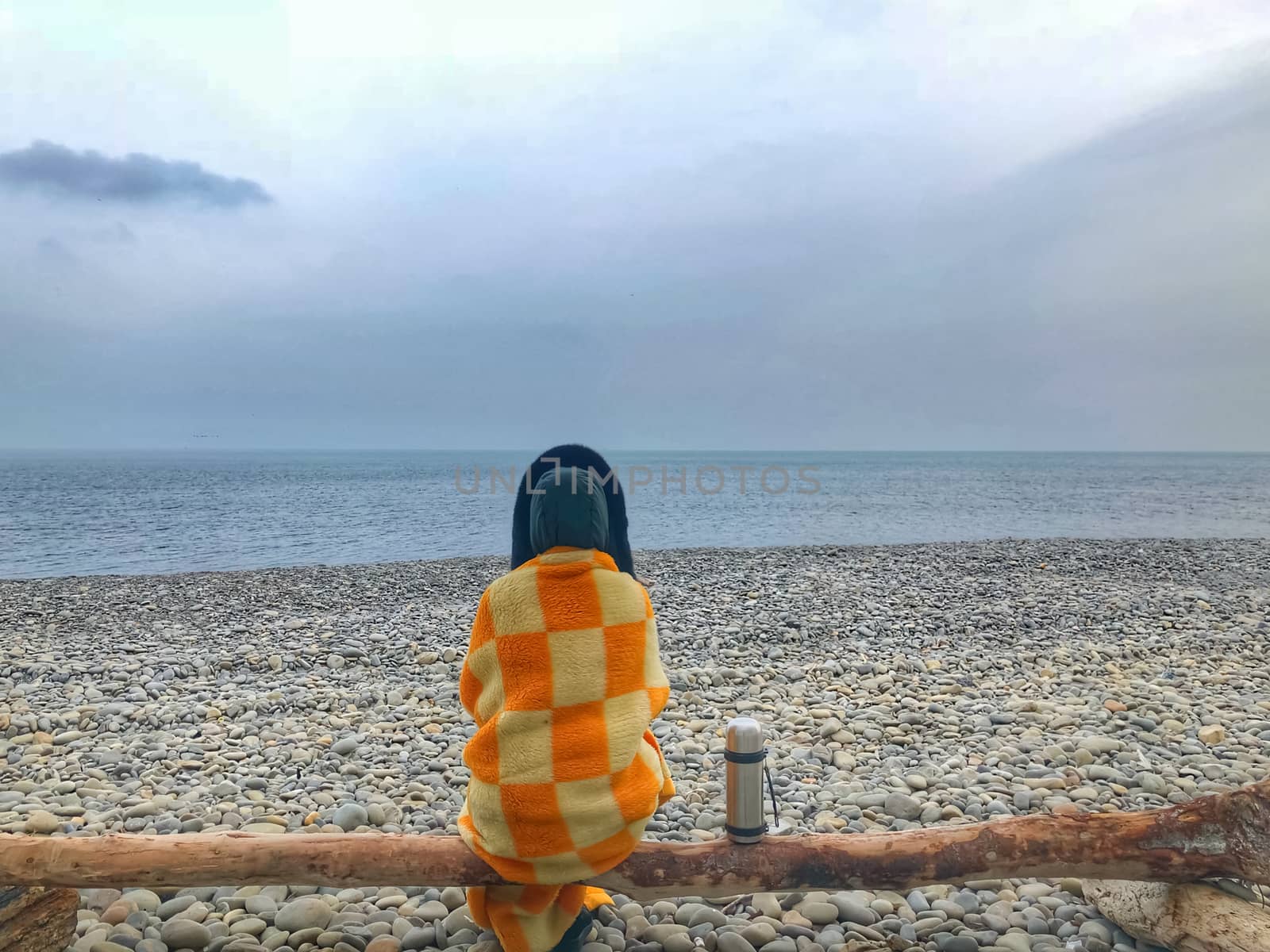 A lonely woman sits by the sea on a log, wrapped in a plaid, next to stands a thermos with tea.