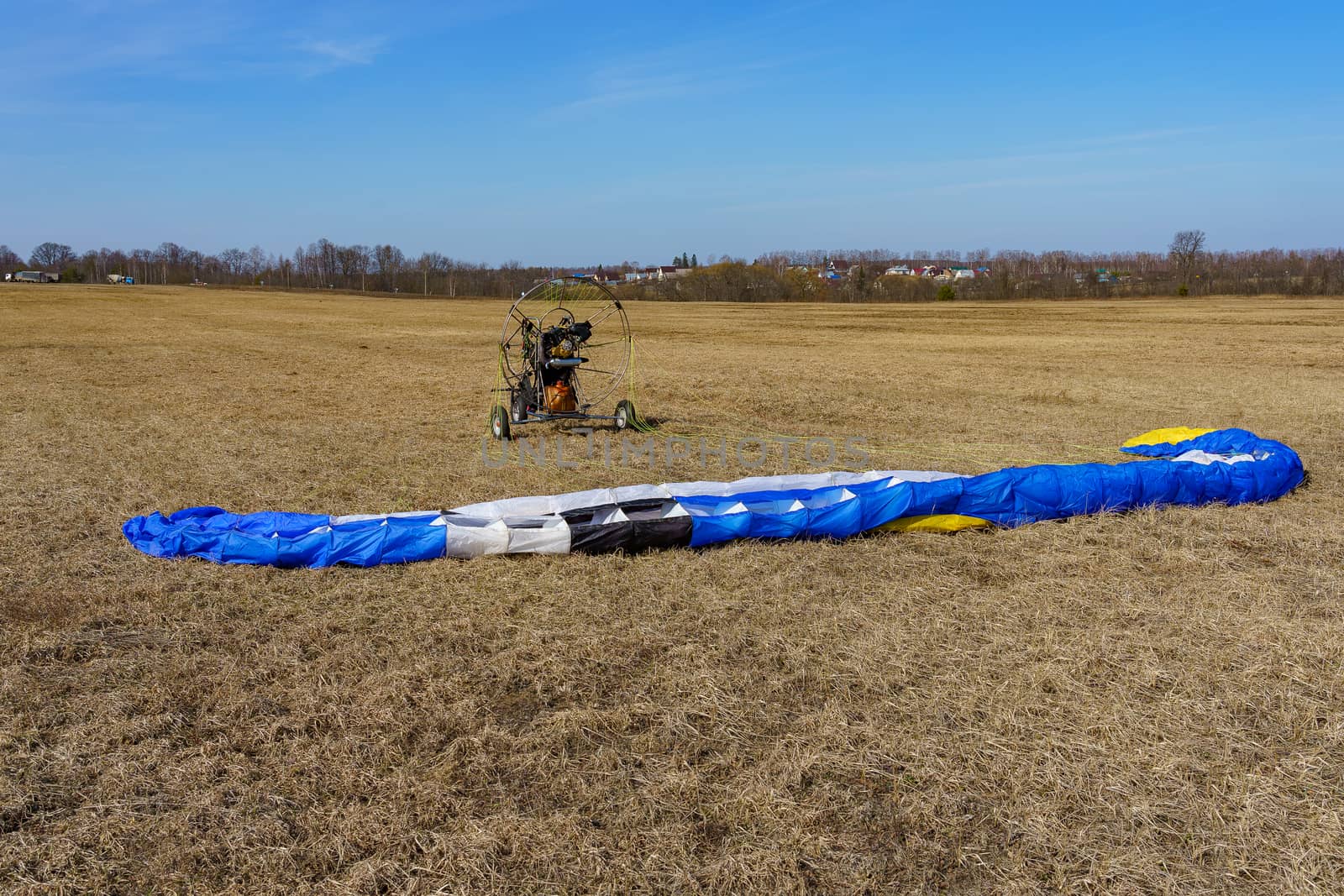 the parachute is spread on the dry grass on the field on a sunny day, in the background a paraglider