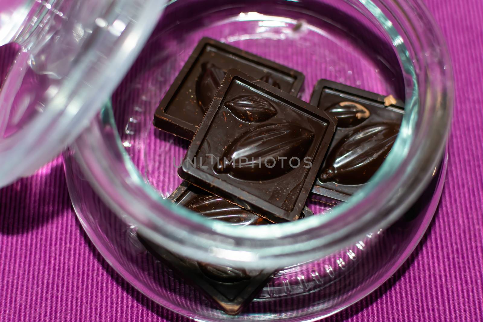 Pieces of handmade chocolate are in a glass jar on a lilac background, close-up, selective focus. Useful and delicious sweets.