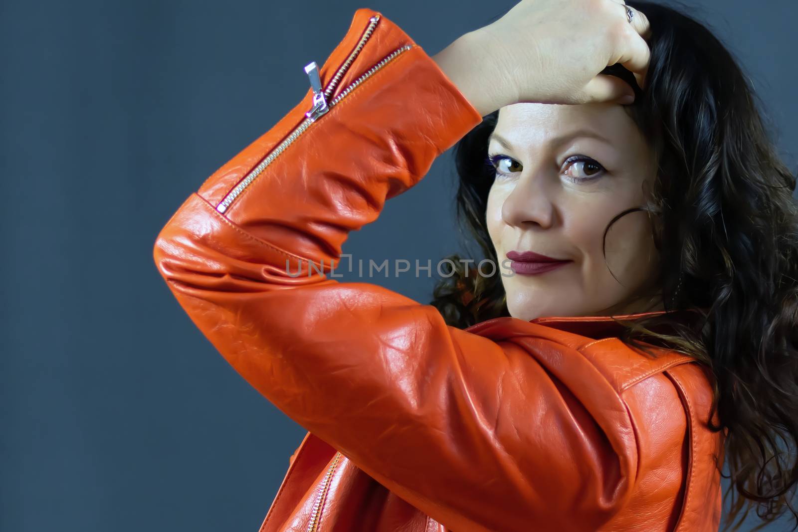 Portrait of a fashionable middle-aged woman in a bright orange leather jacket, posing on a dark gray background by bonilook