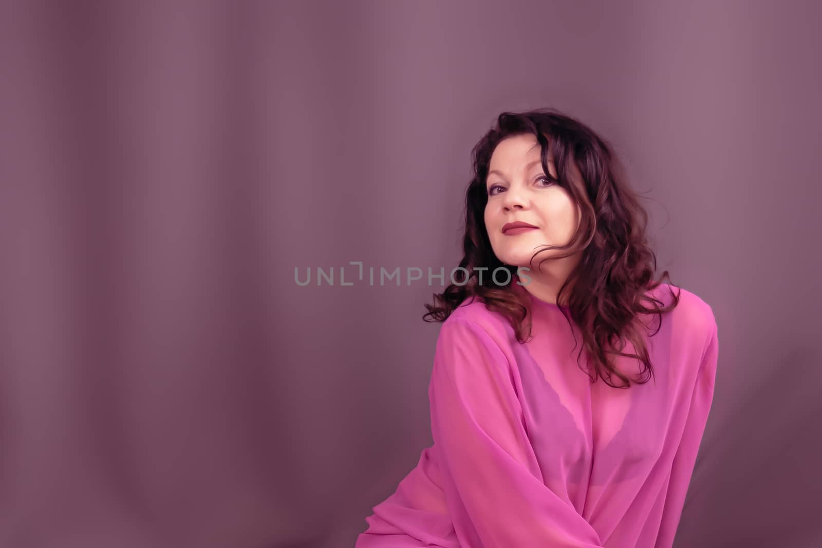 Portrait of a beautiful fashionable middle-aged woman in a bright pink semi-transparent long dress, posing on a dark gray background. The concept of preserving youth at any age.