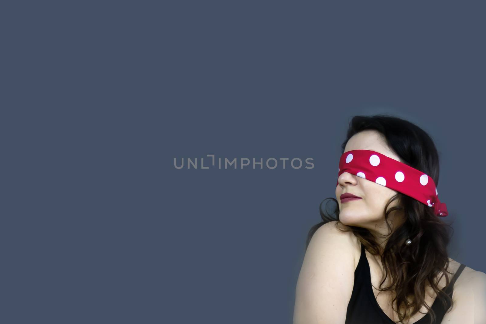 Portrait of a beautiful, fashionable middle-aged woman with long dark hair and a red blindfold covering her eyes, posing against a dark gray background. by bonilook