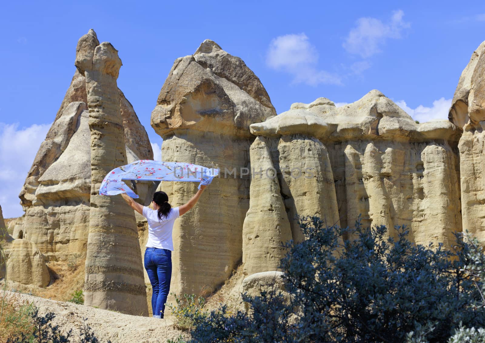 A young woman stands in the wind between huge, old, conical and weathered rocks in the honey valley of Cappadocia under the bright day sun. by Sergii