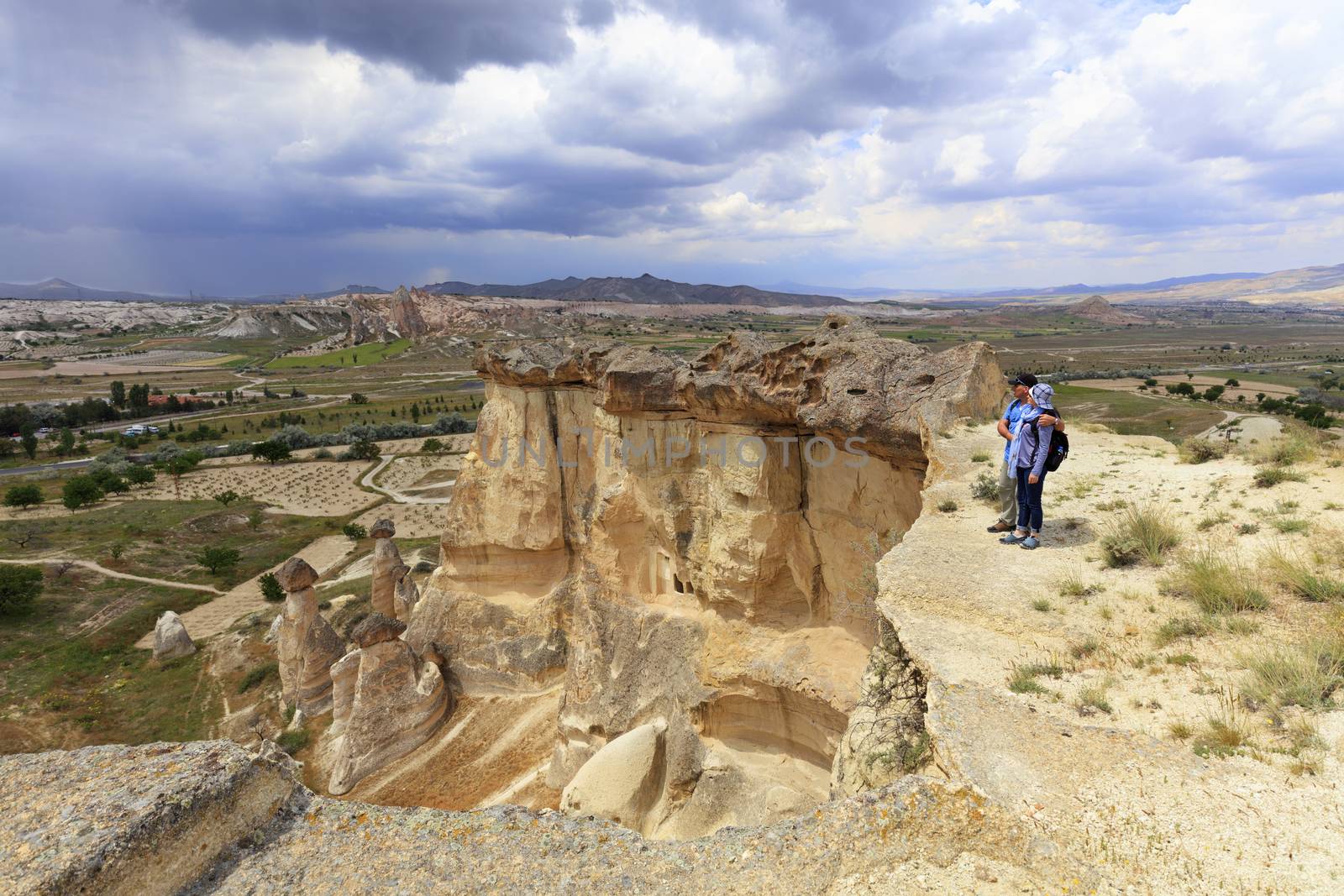 A young couple of tourists with backpacks on their backs stands on the edge of a cliff in Cappadocia and admires the surrounding space against the background of a blue stormy sky and mountain scenery.