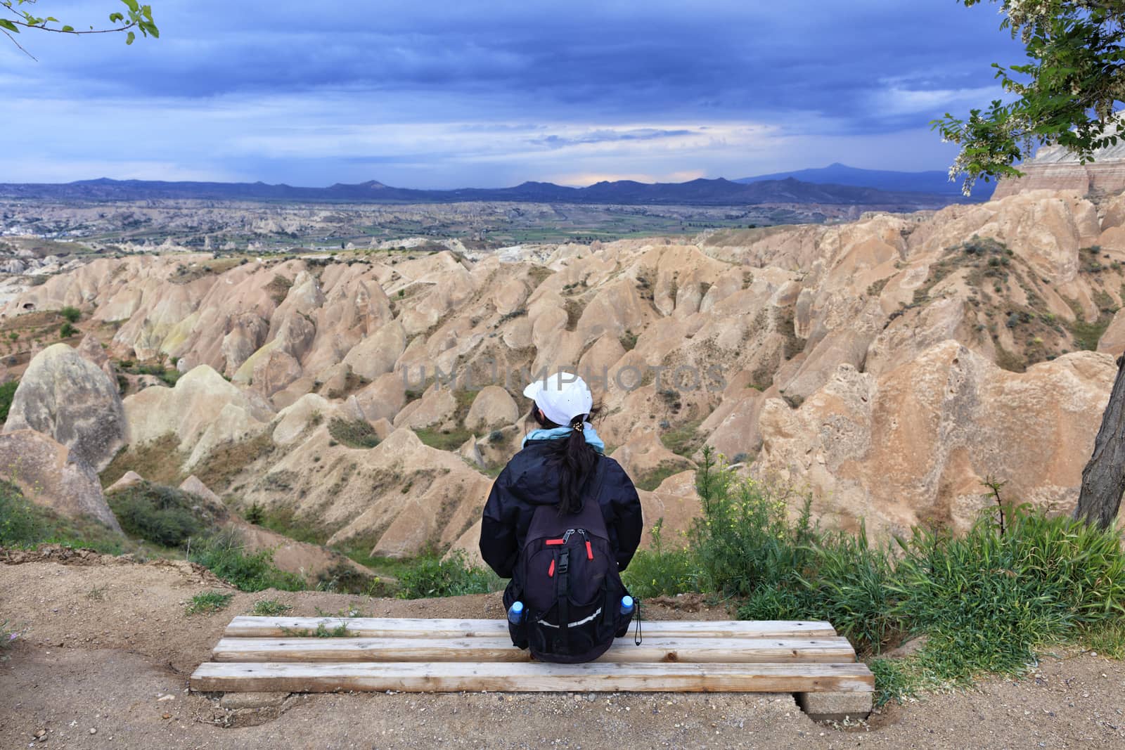 A young girl sits on a wooden bench and looks at the horizon, the mountain ranges of Cappadocia and a stormy sky. by Sergii