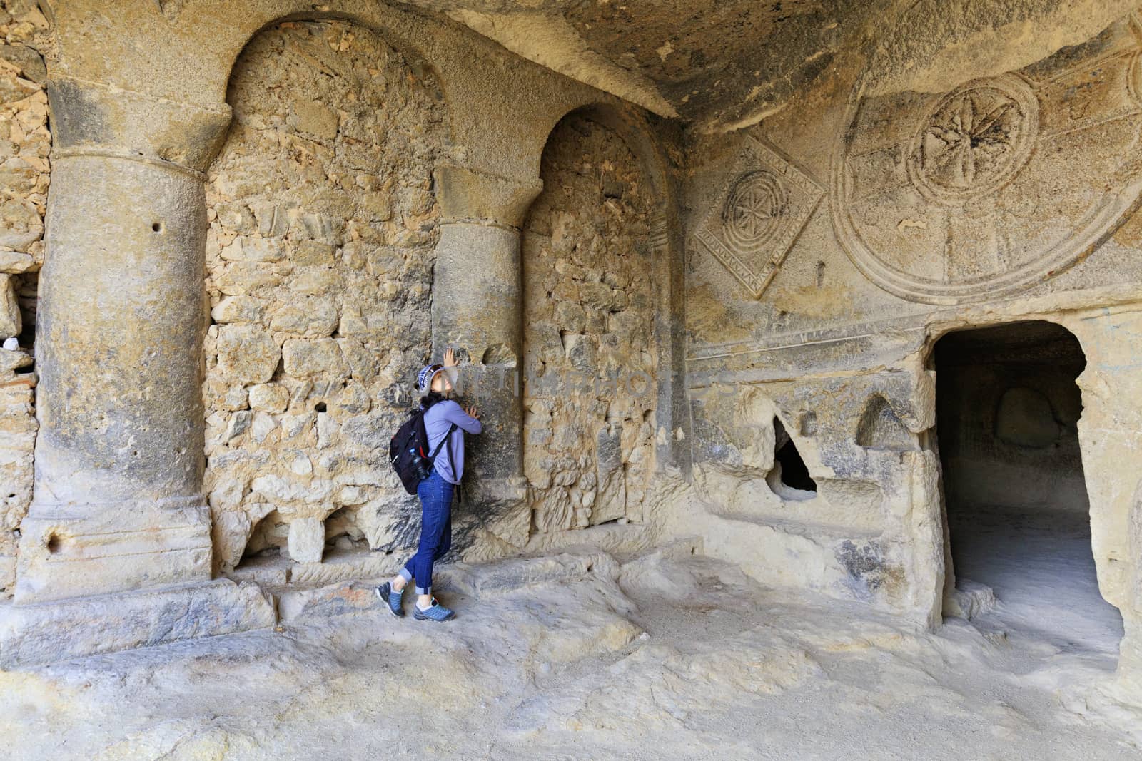 A young woman examines the interior of the old underground church's column hall, carved into a sandstone cliff. by Sergii