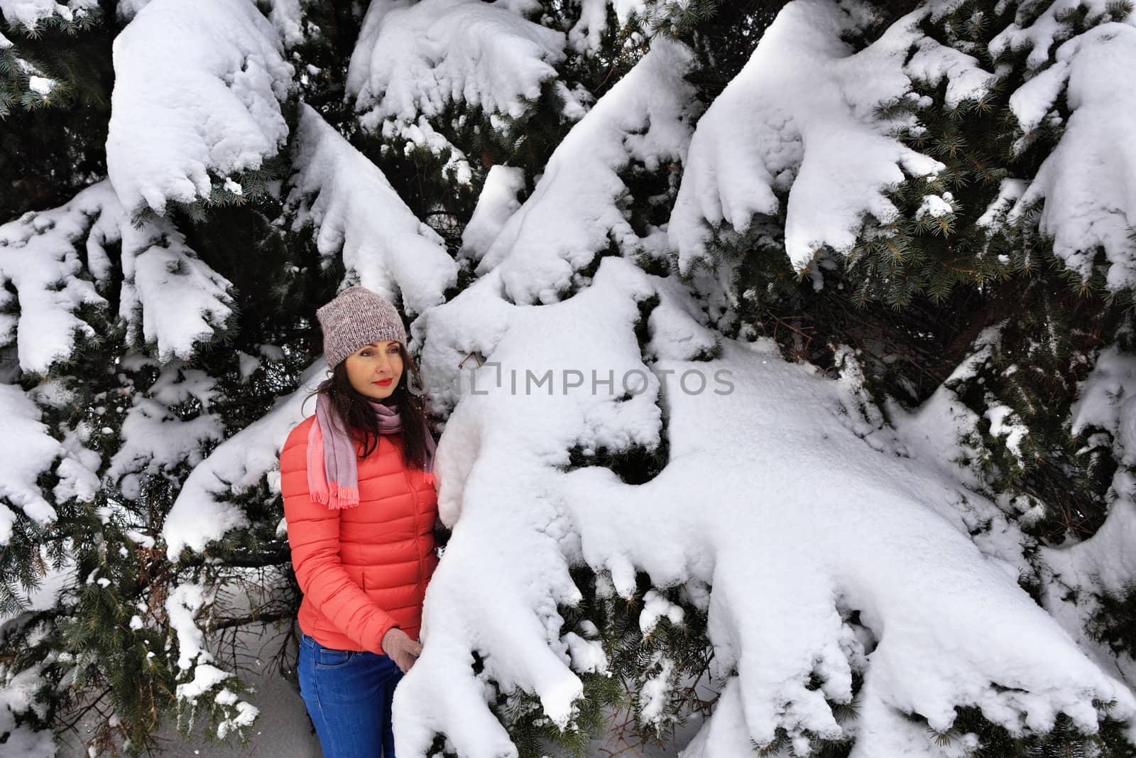 Waist shot of a happy and stylish beautiful woman in a bright coral jacket and blue jeans, smiling, turning her body and head, looking to the right with a carefree facial expression, and enjoying a winter day on the right side of copy space against a winter snow-covered spruce.