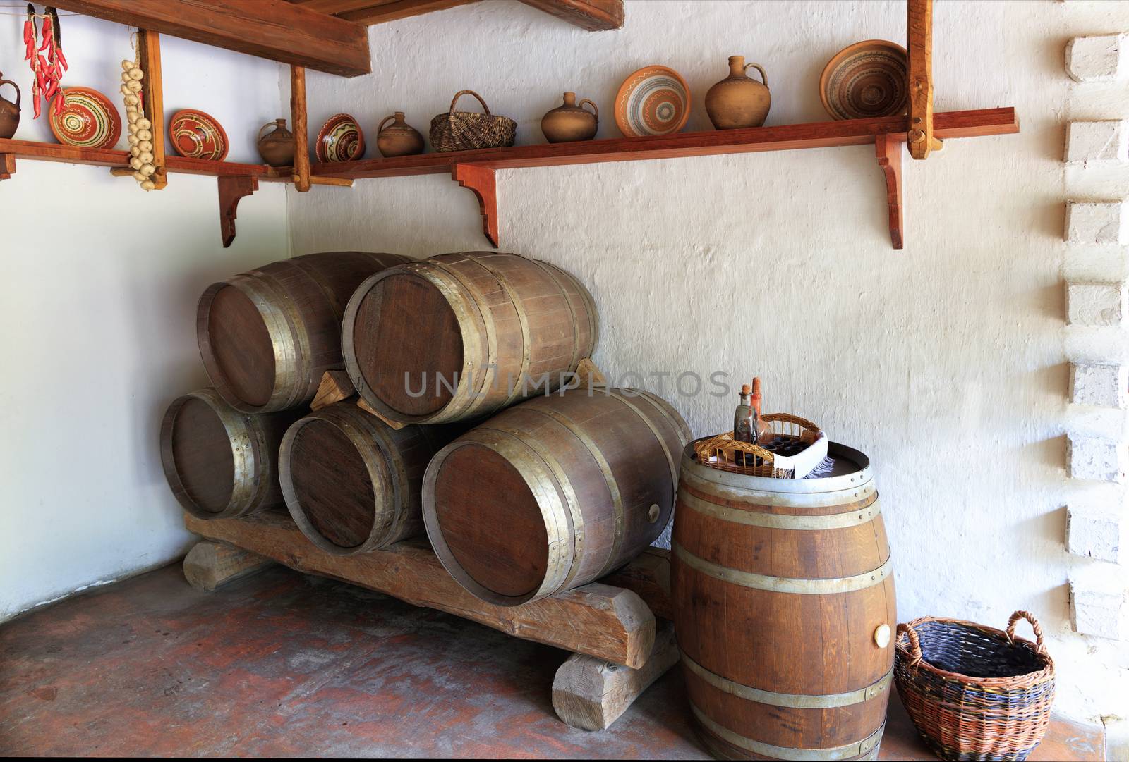 Vintage wooden wine barrels, ceramic painted bowls, plates and jugs on a wooden shelf on the wall, a wicker basket with glassware in the interior of an old Ukrainian rural home.