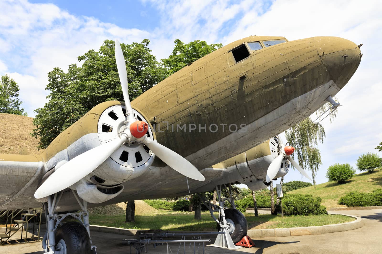 An old transport military aircraft Lisunov Li-2 of the Second World War, stands in a park against a cloudy blue sky.
