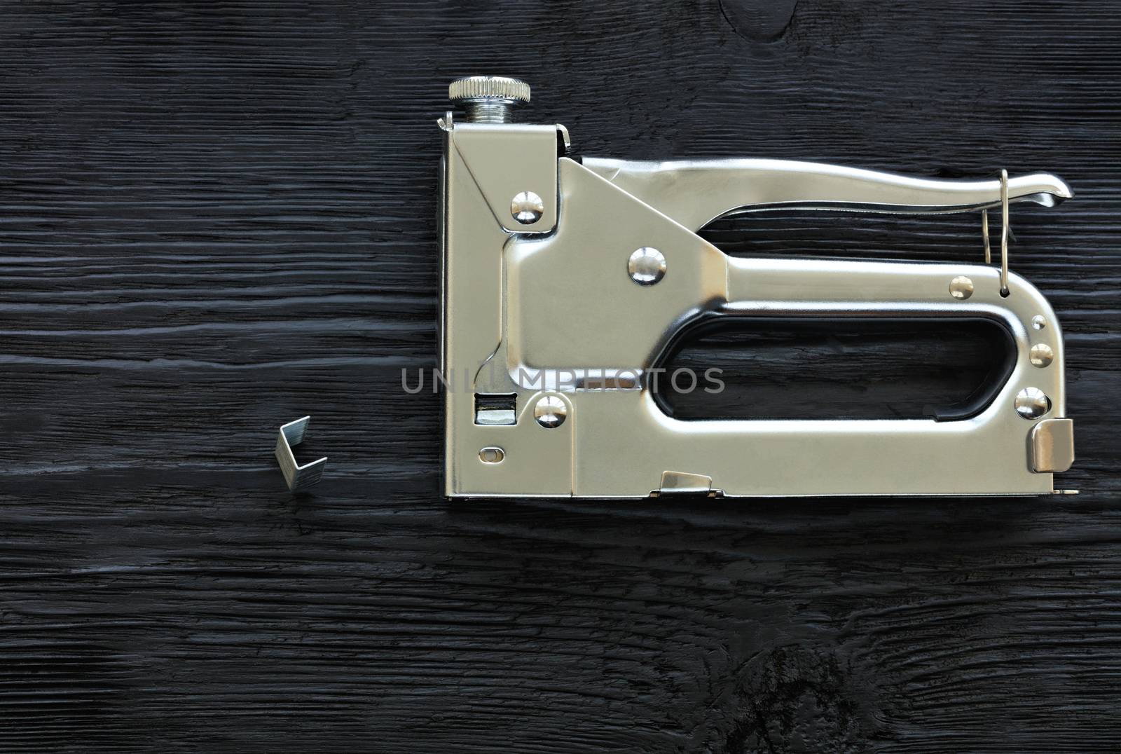 Metal industrial chrome stapler and brackets to it lie on a black wooden table with the texture of old wood.