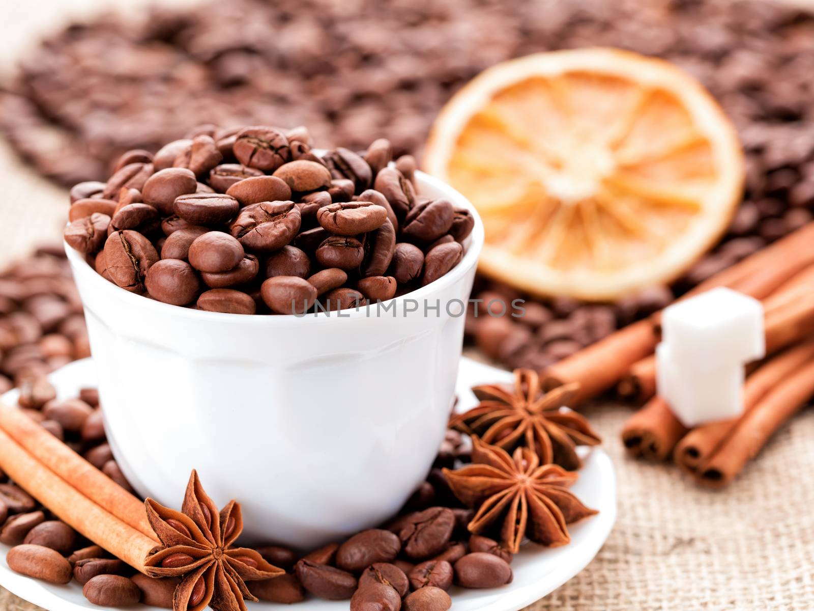 Grains of black coffee in a white porcelain dish. Cinnamon sticks and star anise on a white porcelain dish. Dried lemon slices and sugar in the background.