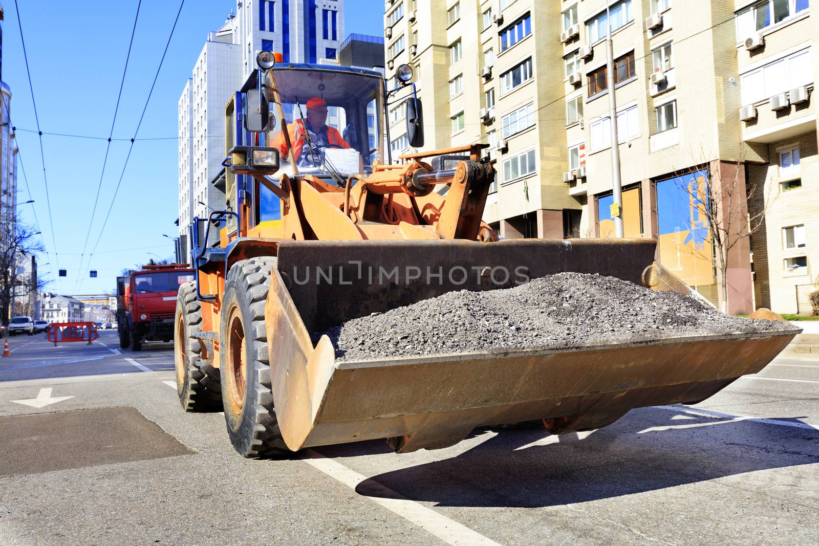 A road worker carries gravel in a heavy construction bulldozer bucket during roadway repair. by Sergii